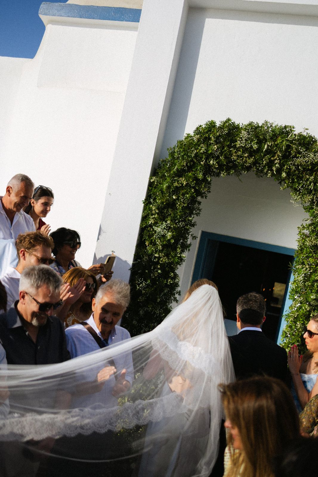 Bride walking into a whitewashed chapel on Kimolos under a green arch, her veil caught in the wind — wedding photographed by John Xenos
