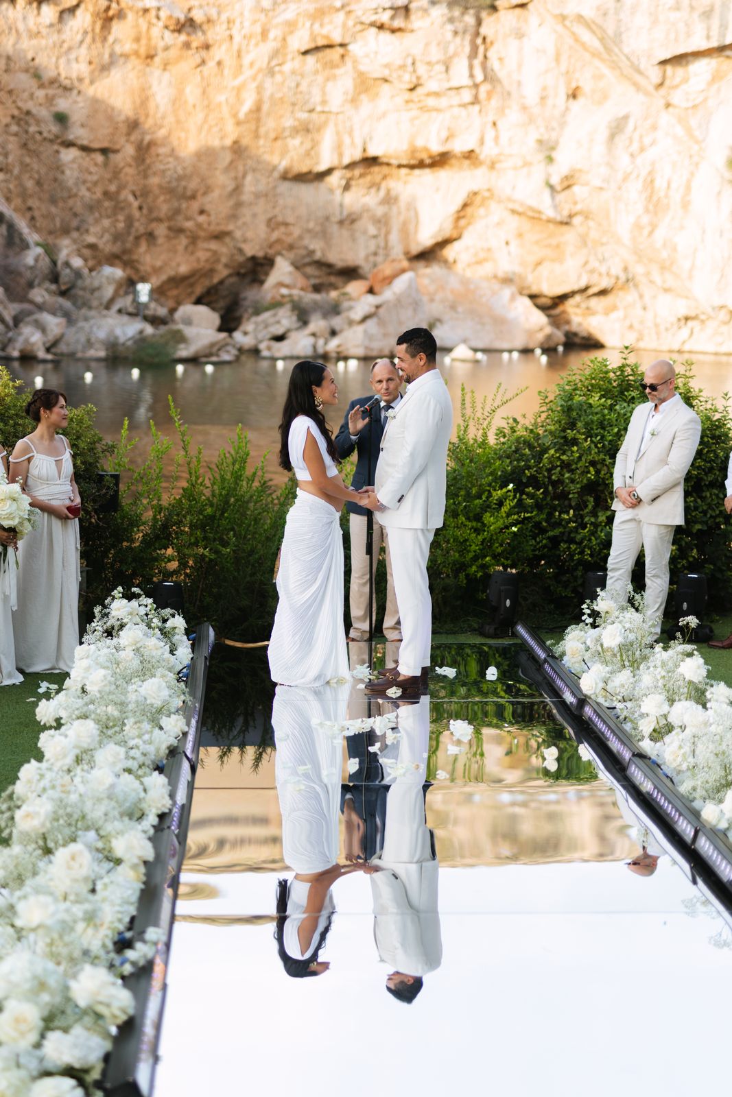 Bride and groom holding hands before the officiant, reflected in the mirror aisle at Lake Vouliagmeni
