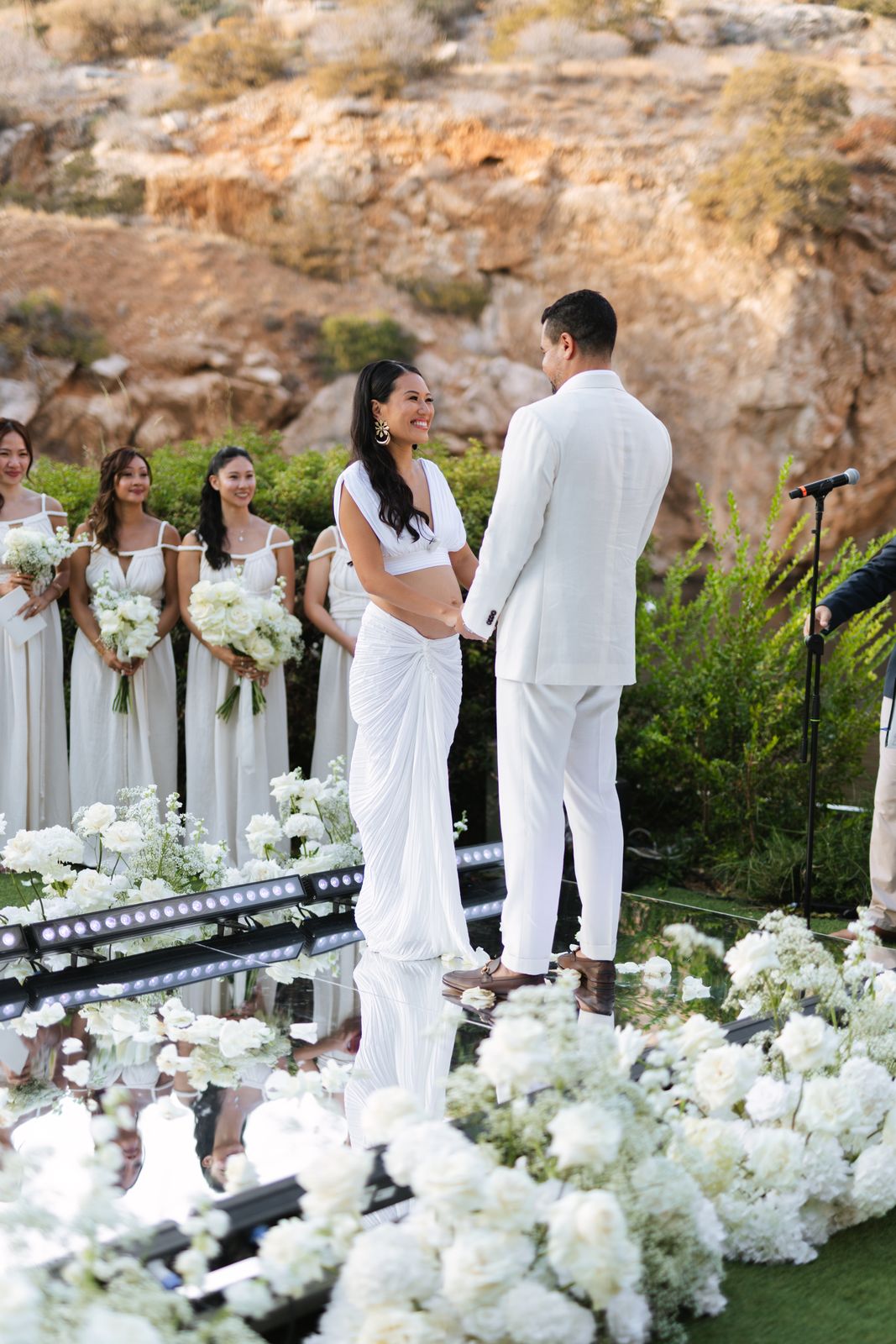 Couple holding hands at the altar as bridesmaids hold white bouquets, cliff glowing at Lake Vouliagmeni