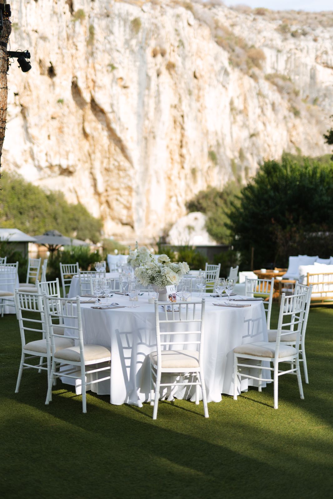 Dinner tables with white chairs on the lawn beneath the cliff at Lake Vouliagmeni