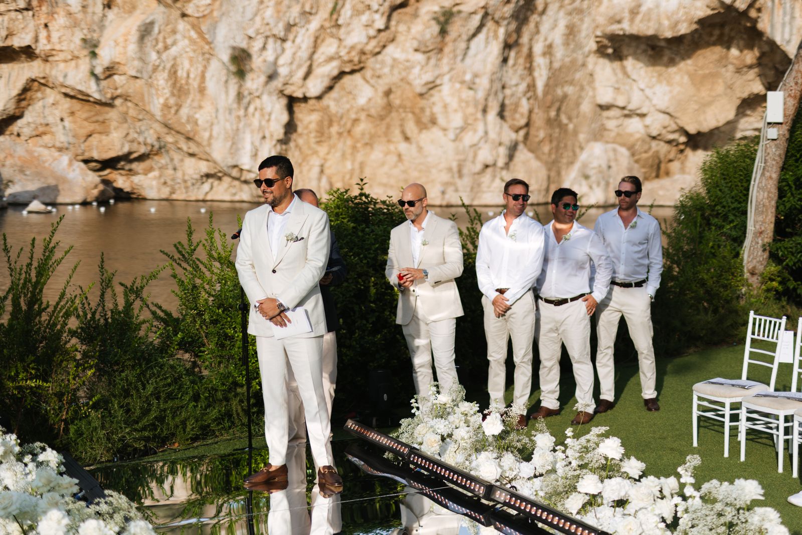 Groom and groomsmen in white waiting at the end of the mirror aisle, Lake Vouliagmeni behind
