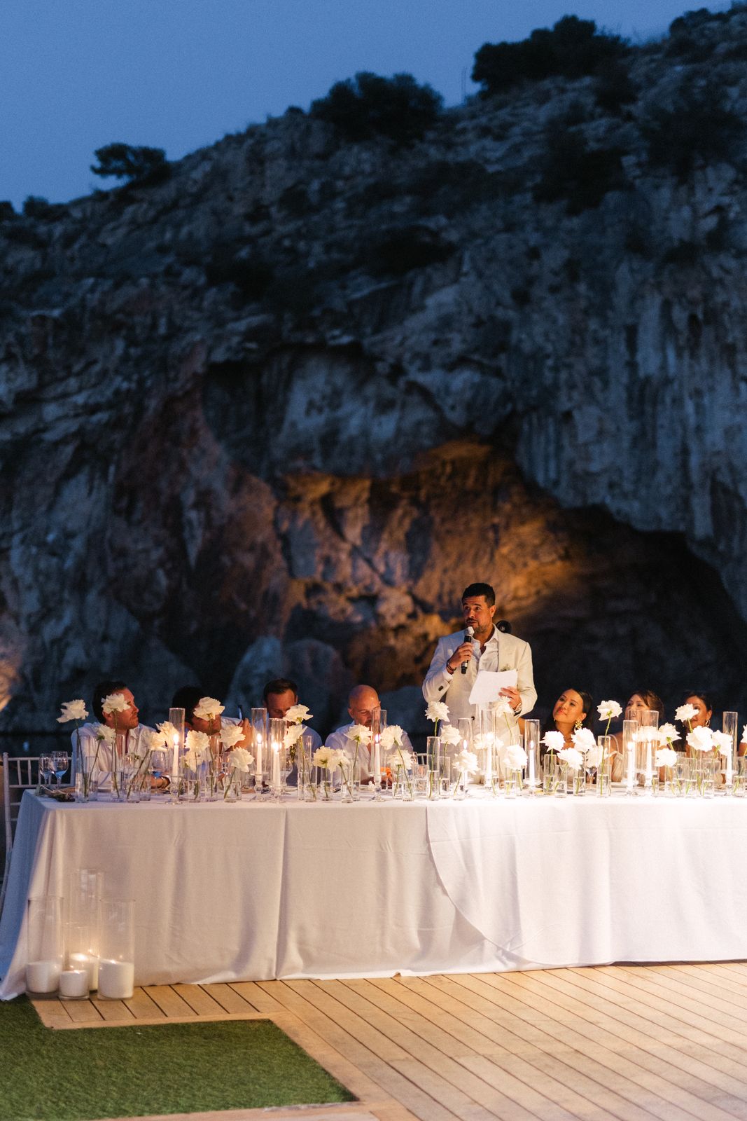 Groom's speech at the candlelit head table under the floodlit cliff, Lake Vouliagmeni at dusk