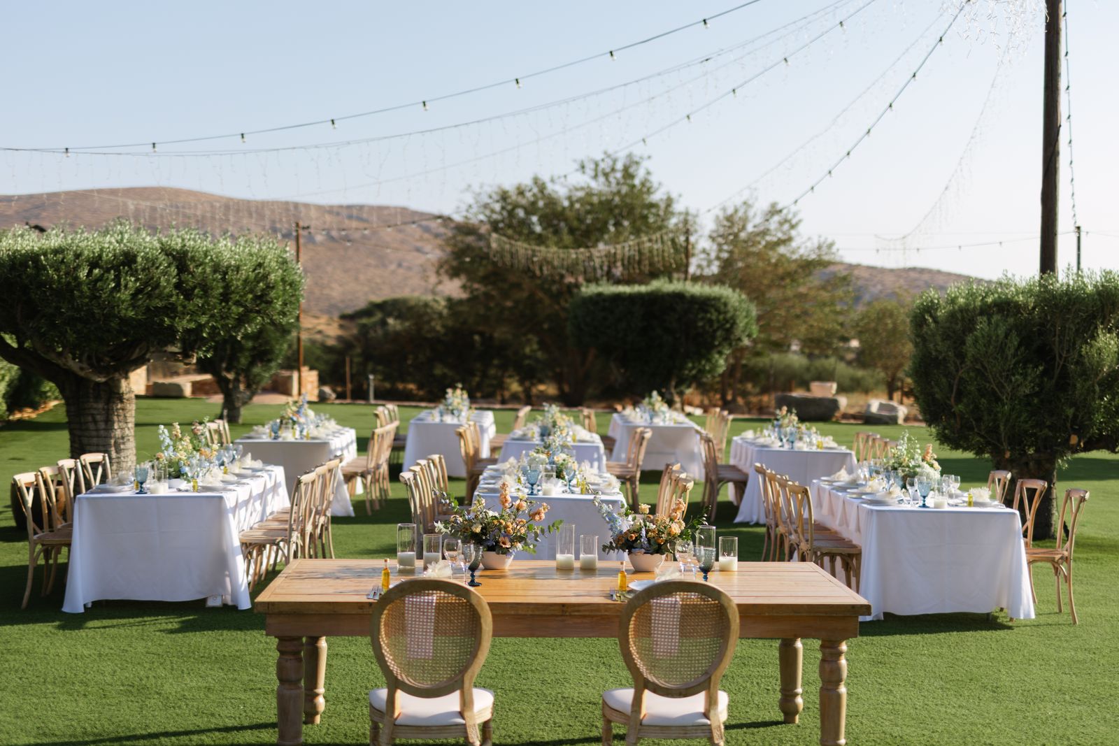 Dinner tables set on the Pyrgos Melissourgou lawn under string lights, Attica hills behind